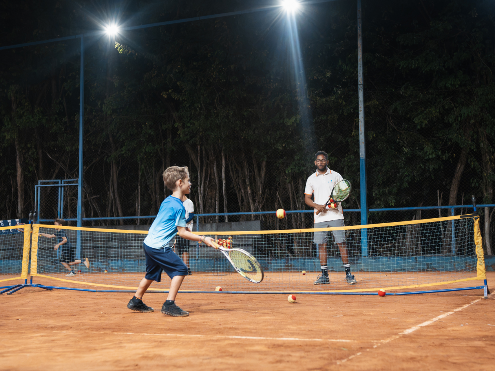 Criança em treino noturno na quadra de saibro com o professor